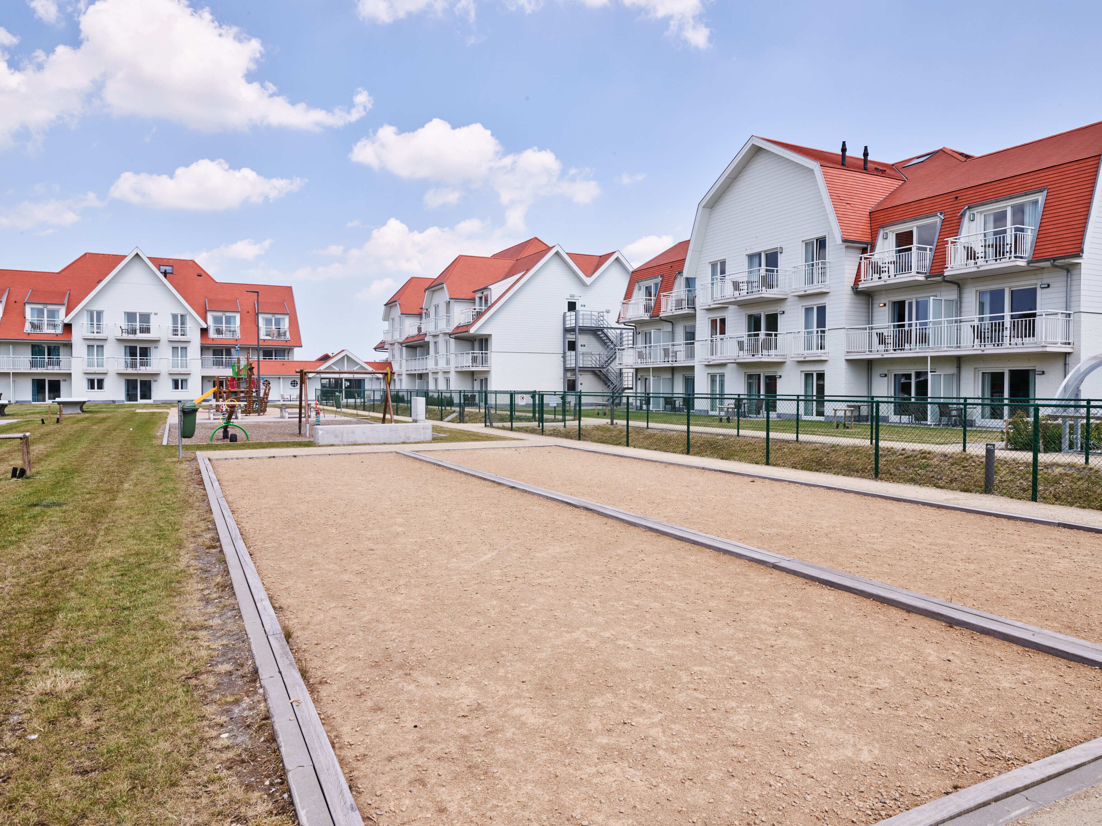 A row of white buildings with red roofs, a playground, and a sandy area.