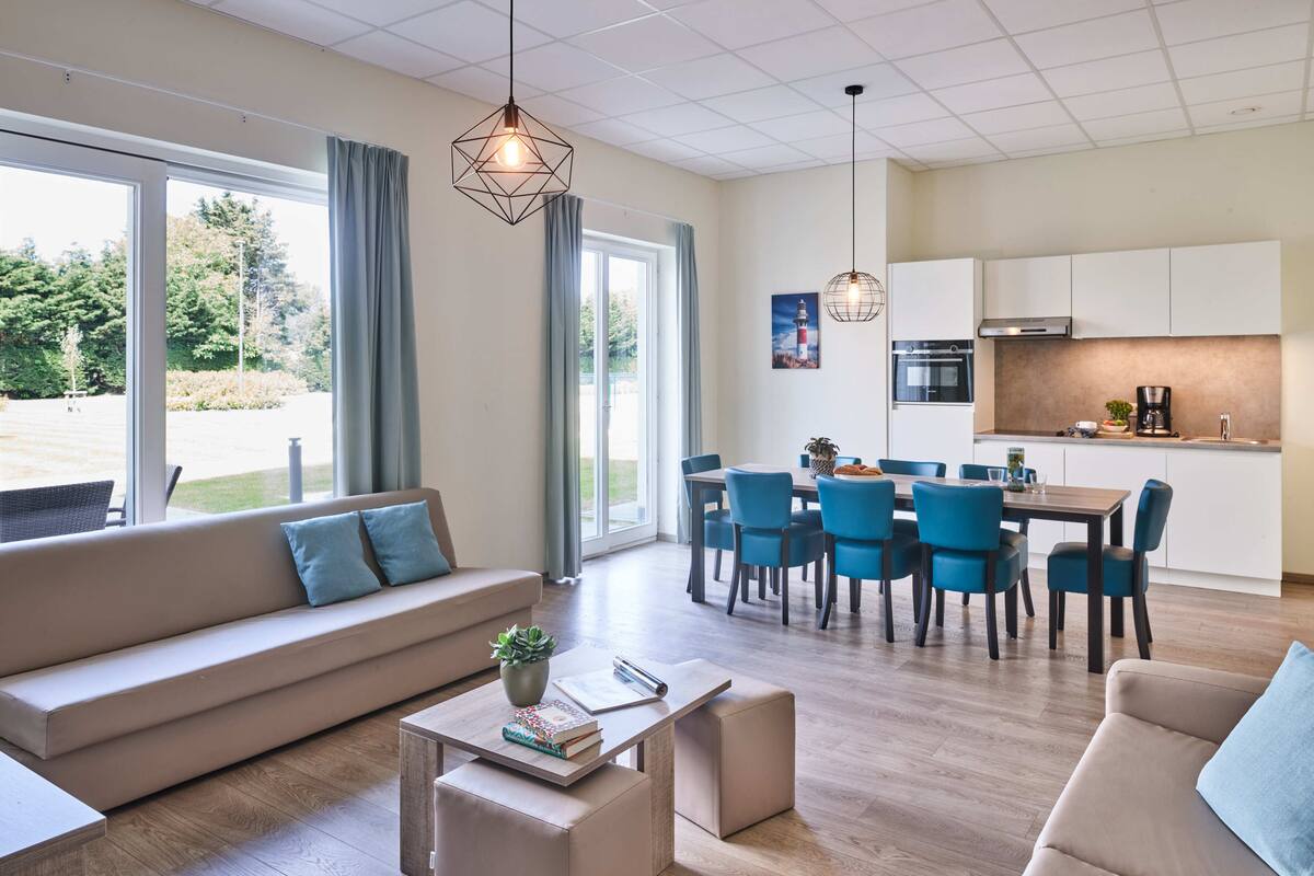 Una sala moderna con sofá, mesa de centro y comedor con sillas. Se observa una cocina con armarios y electrodomésticos.