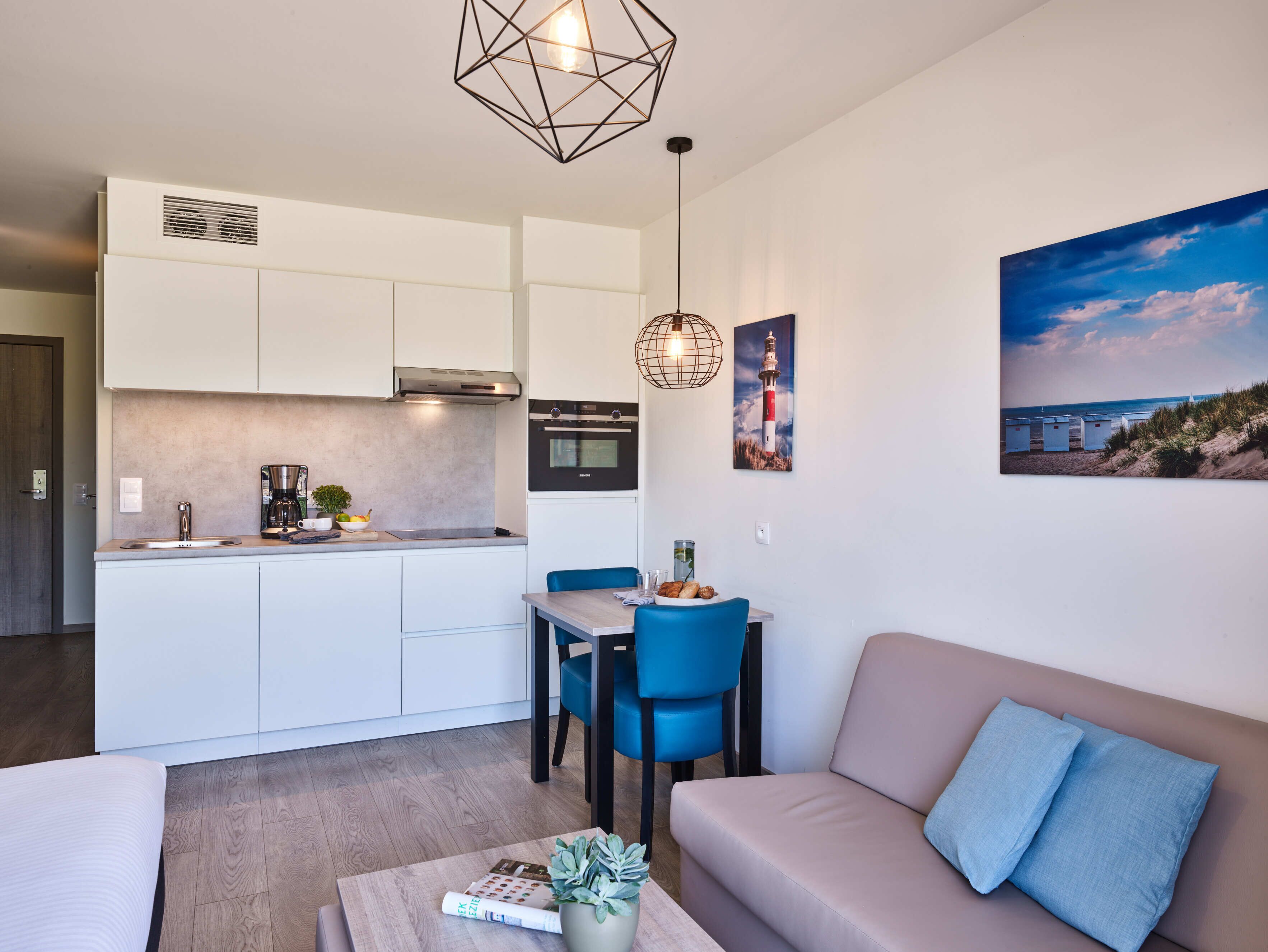 Una cocina moderna con gabinetes blancos, horno empotrado y cafetera. Hay un comedor con una silla azul y una mesa, y una sala de estar con sofá color beige y un cojín azul.