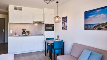 Una cocina moderna con gabinetes blancos, horno empotrado y cafetera. Hay un comedor con una silla azul y una mesa, y una sala de estar con sofá color beige y un cojín azul.