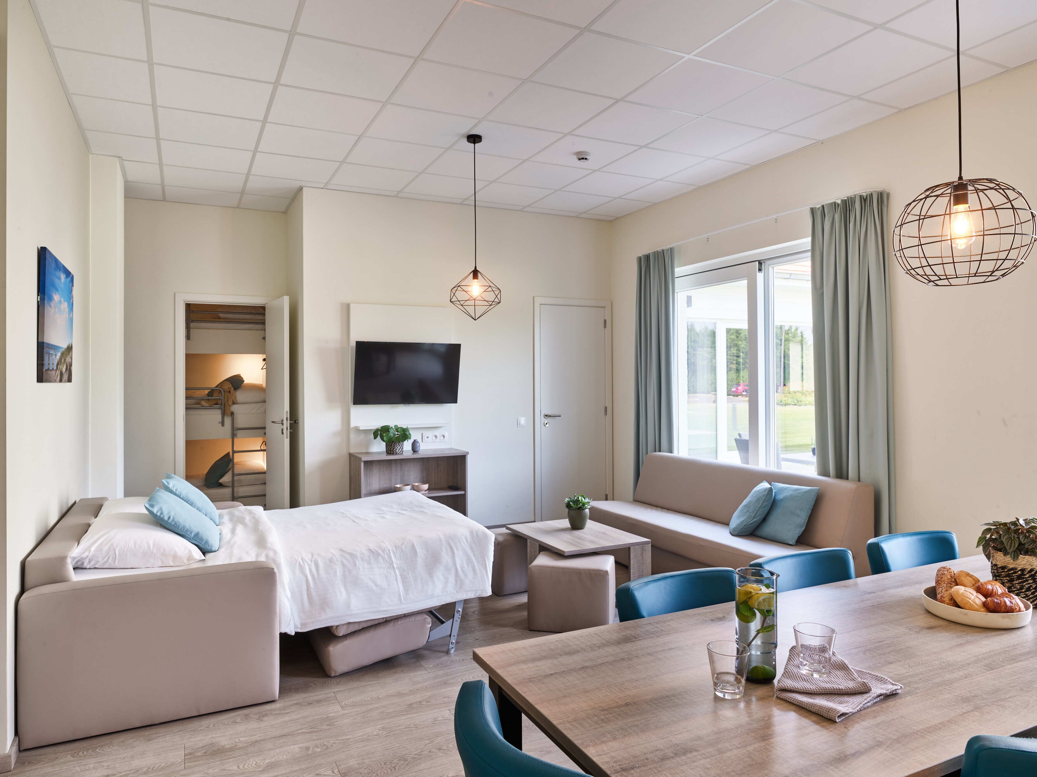 A modern hotel room with a bunk bed, a sofa, a dining table, and a TV.