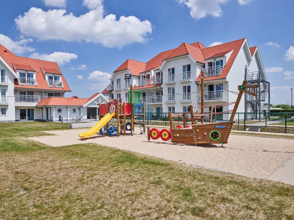 A playground with a pirate ship play structure and a yellow slide, set against a backdrop of white buildings with red roofs. - Holiday Suites Nieuwpoort (Nieuwpoort)