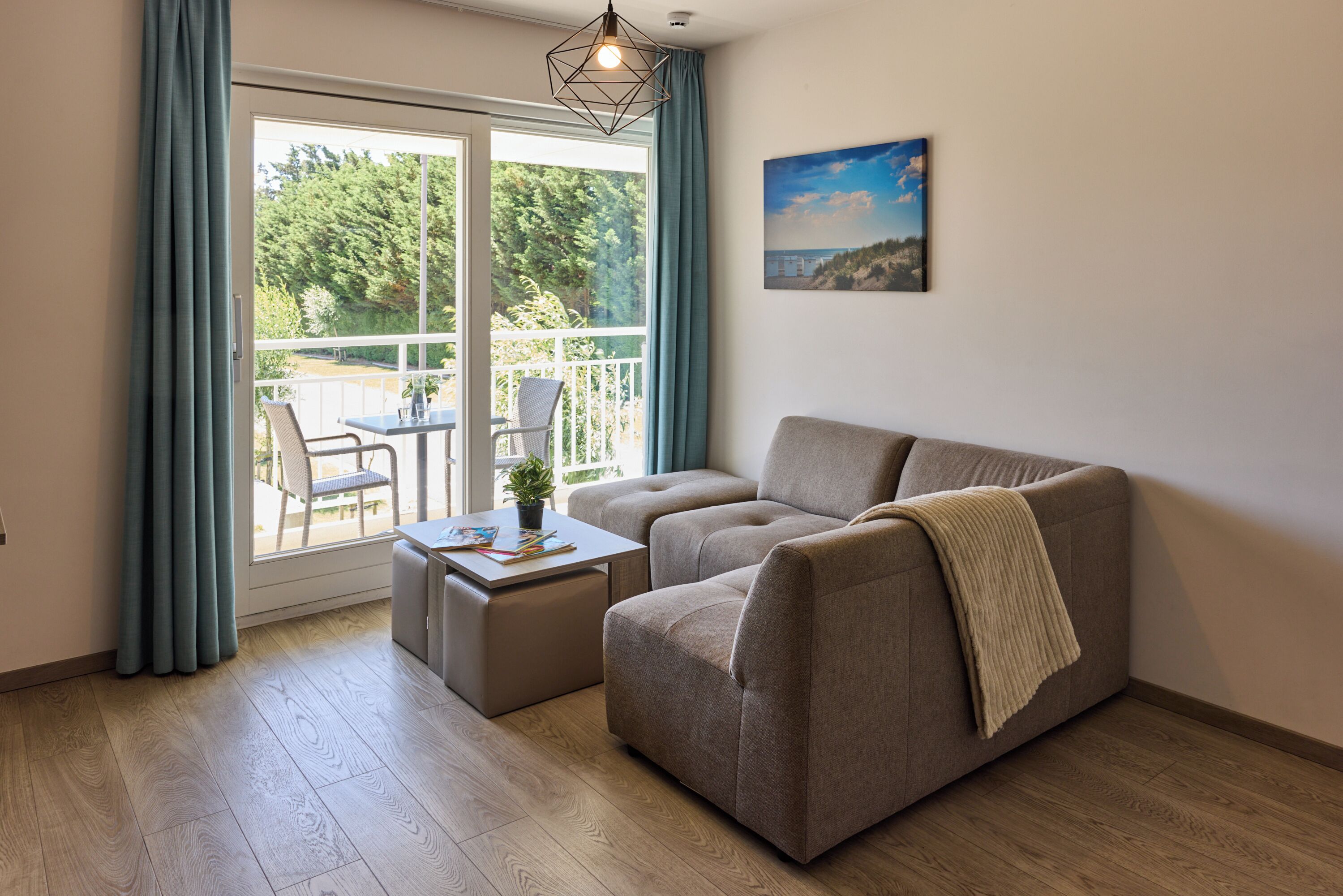Una sala de estar con un sofá, una mesita y una silla. Tiene un balcón con vistas a árboles y un edificio.