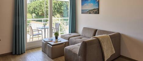 A living room with a sofa, a small table, and a chair. There is a balcony with a view of trees and a building.