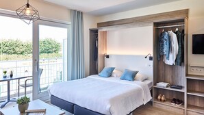 A modern bedroom with a large bed, a bedside table with a plant, a built-in wardrobe, and a balcony with a view. - Holiday Suites Nieuwpoort (Nieuwpoort)