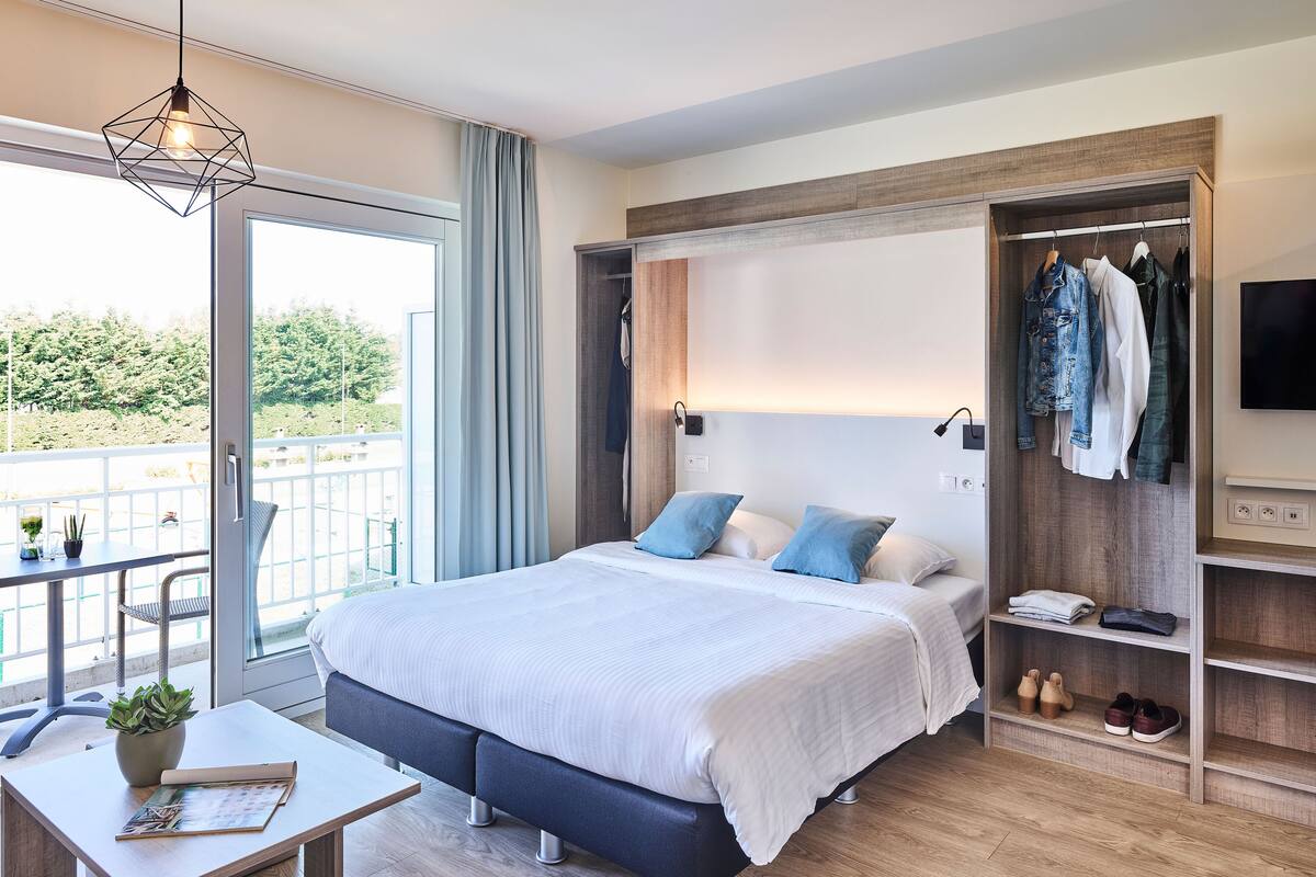 Un dormitorio moderno con una cama grande, una mesita de noche con una planta, un armario empotrado y un balcón con vistas.