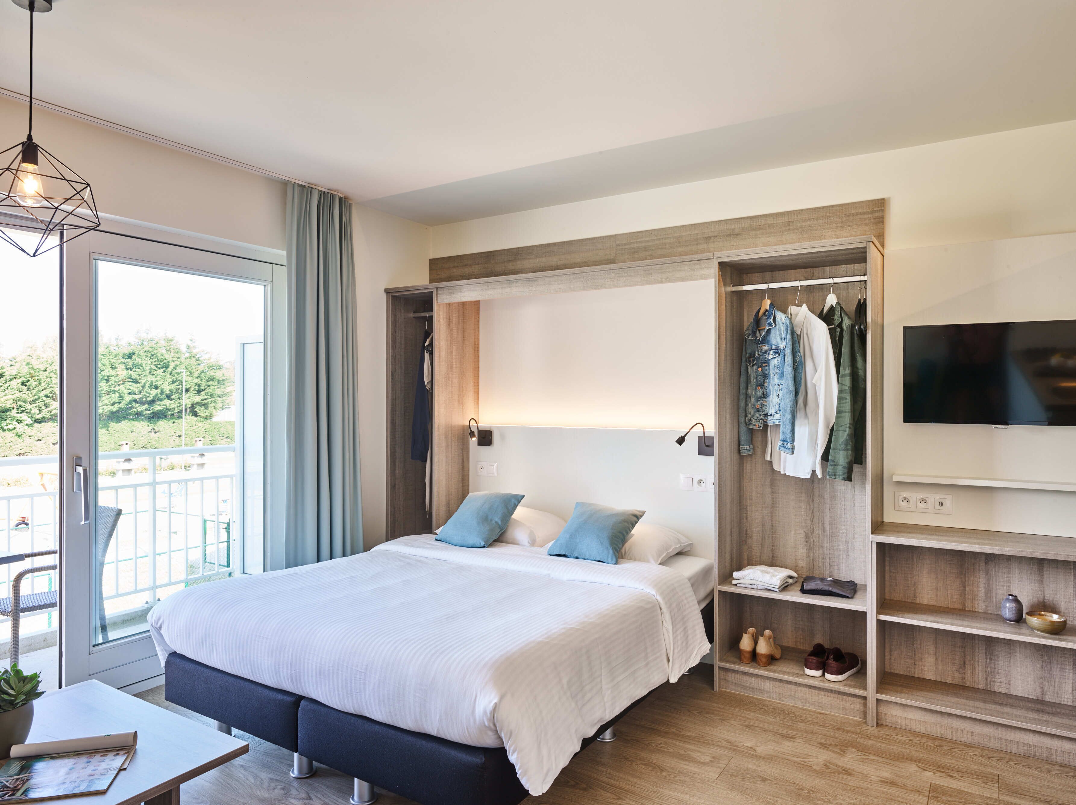 A modern hotel room with a large bed, a TV mounted on the wall, a wardrobe, and a balcony with a view of trees.