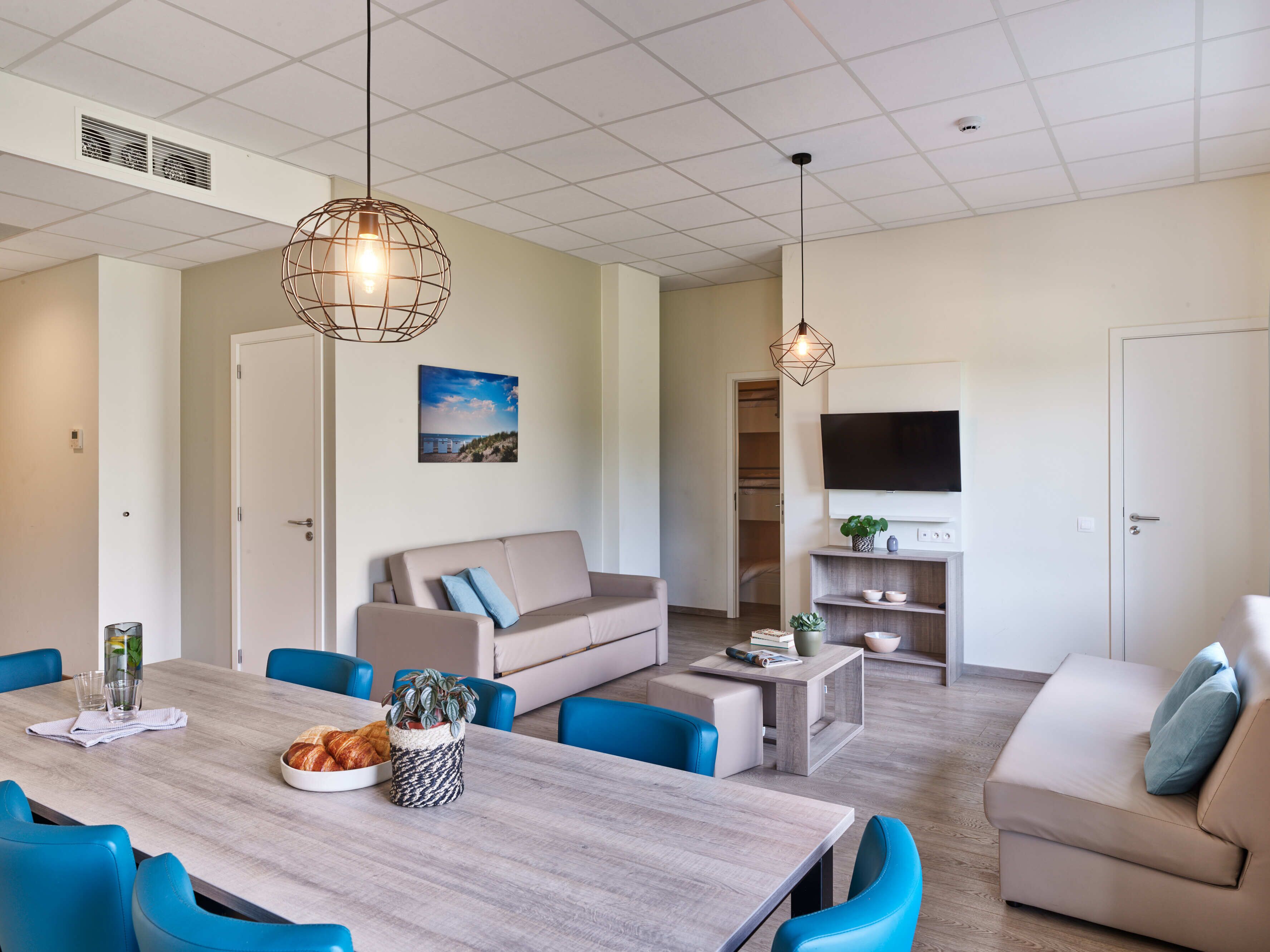A modern living room with a sofa, coffee table, and dining table. There is a TV mounted on the wall and a window with a view.