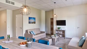 A modern living room with a sofa, coffee table, and dining table. There is a TV mounted on the wall and a window with a view.