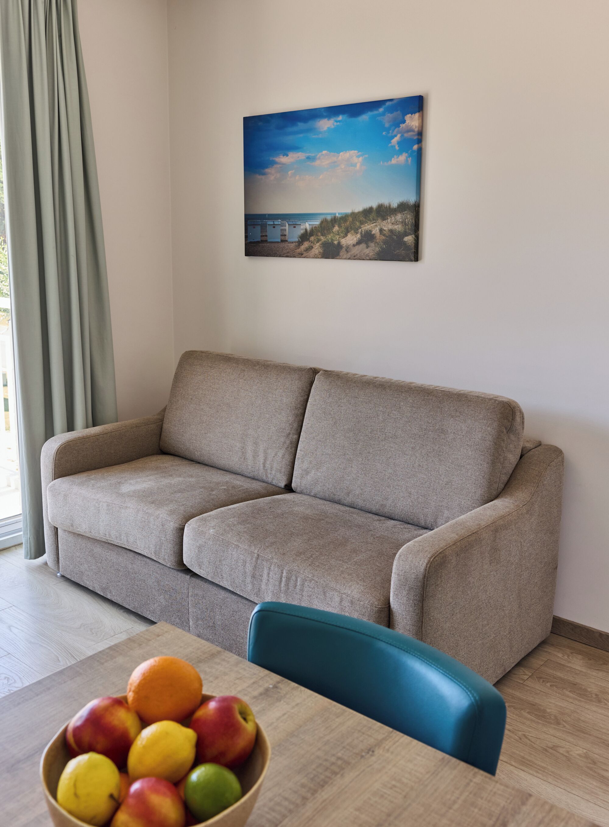 Un canapé beige, une chaise bleue et une table sur laquelle se trouve un saladier rempli de fruits.