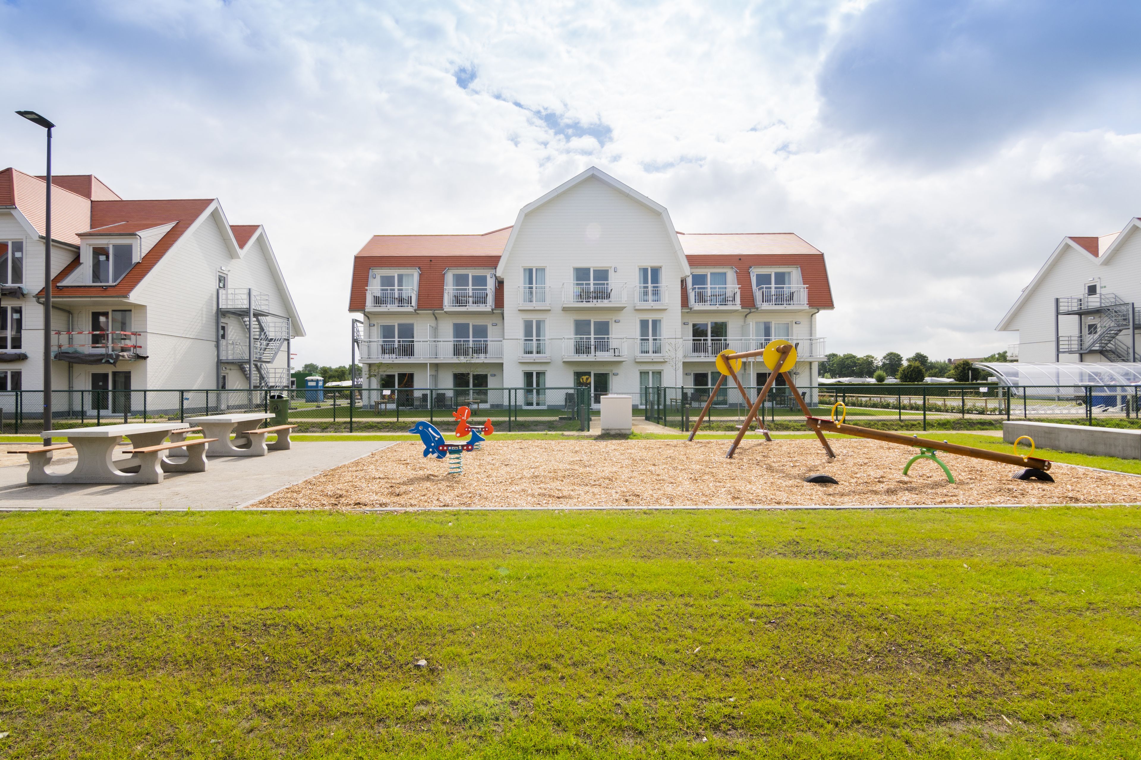 A playground with swings and a seesaw, surrounded by a grassy area and a building with multiple windows and balconies.