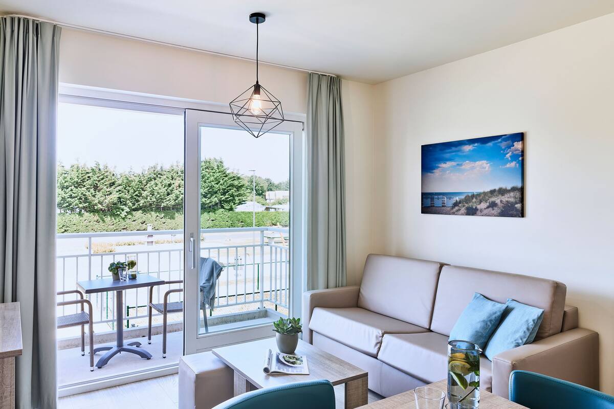 Una sala de estar moderna con sofá, mesa de comedor y balcón con vistas a zonas verdes y una playa.