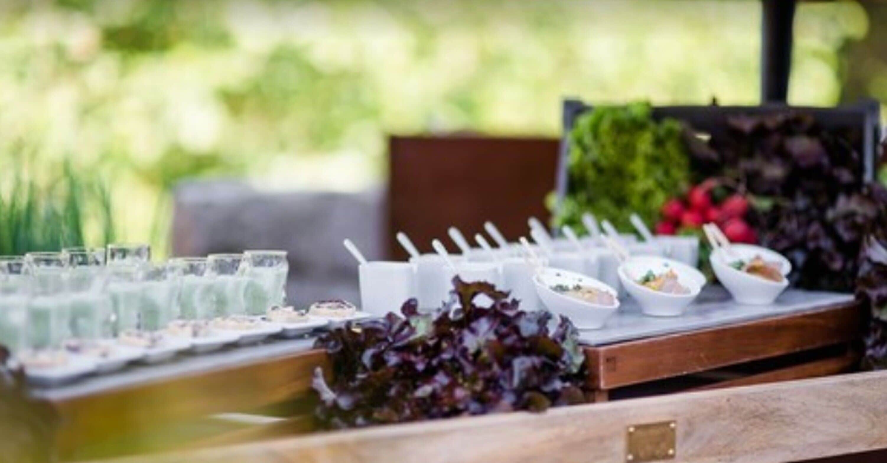 A buffet table with various dishes and drinks, set outdoors with greenery in the background.
