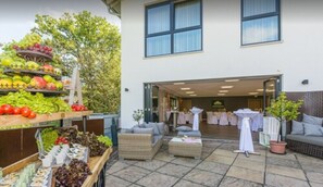 An outdoor patio area with a fruit and vegetable display, wicker furniture, and a view of the building's interior. - Romantik Hotel Ahrenberg (Bad Sooden-Allendorf)