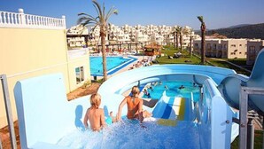 Un tobogán acuático con gente en la base, una zona de piscina y edificios al fondo.