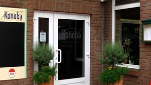 A brick building with a white door labeled "Konoba Restaurant Bistro." There are two large potted plants in front of the door. - Hotel Konoba Restaurant - Bistro (Meckenheim)