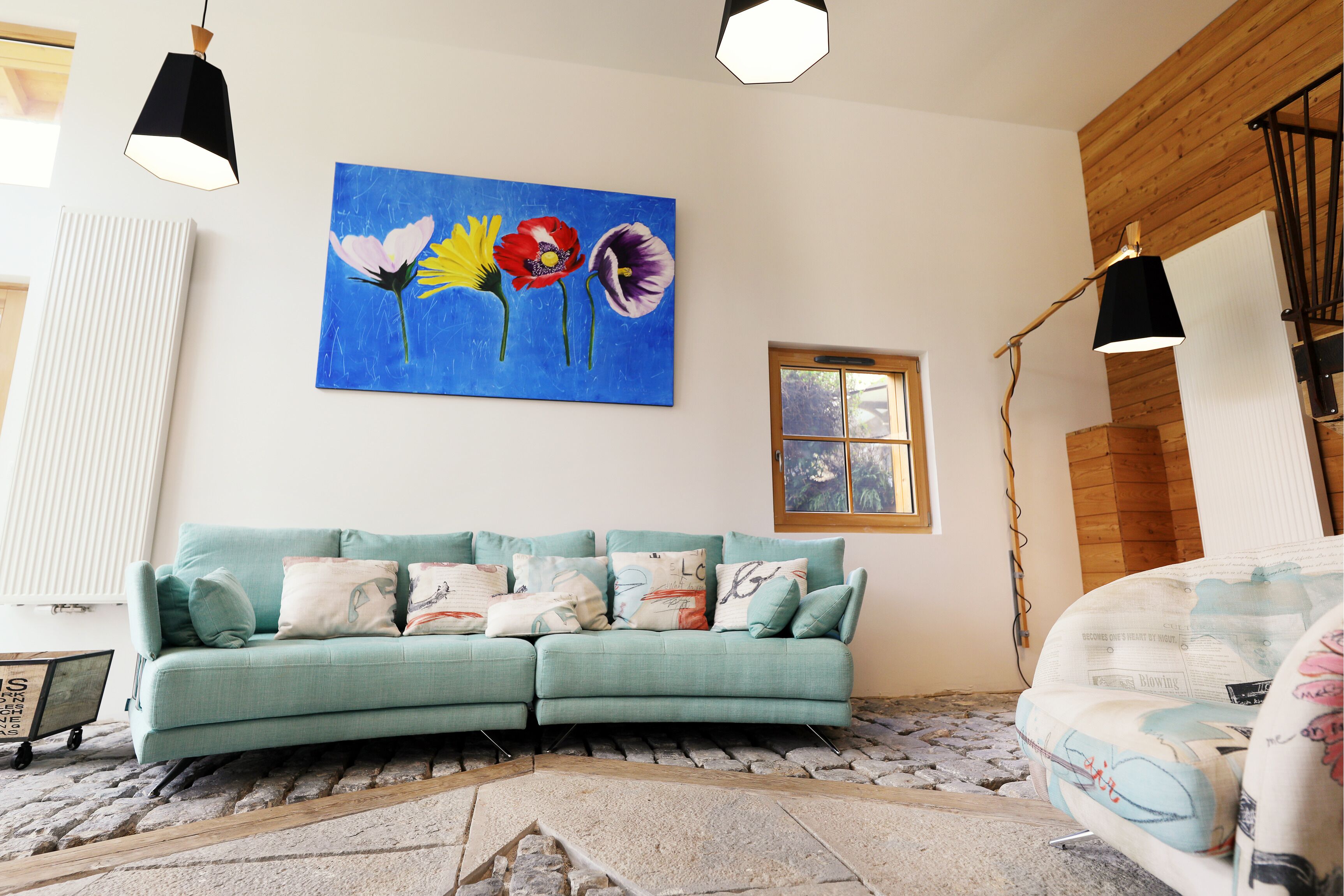 A teal sofa with multiple cushions, a painting of flowers on the wall, a window, and a lamp.