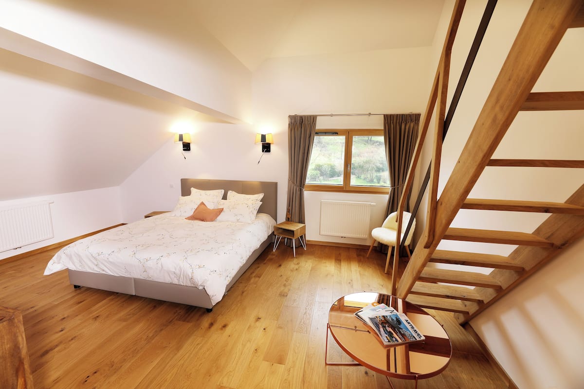 A bedroom with a bed, bedside tables, a window with curtains, a wooden staircase, and a small table with a magazine.