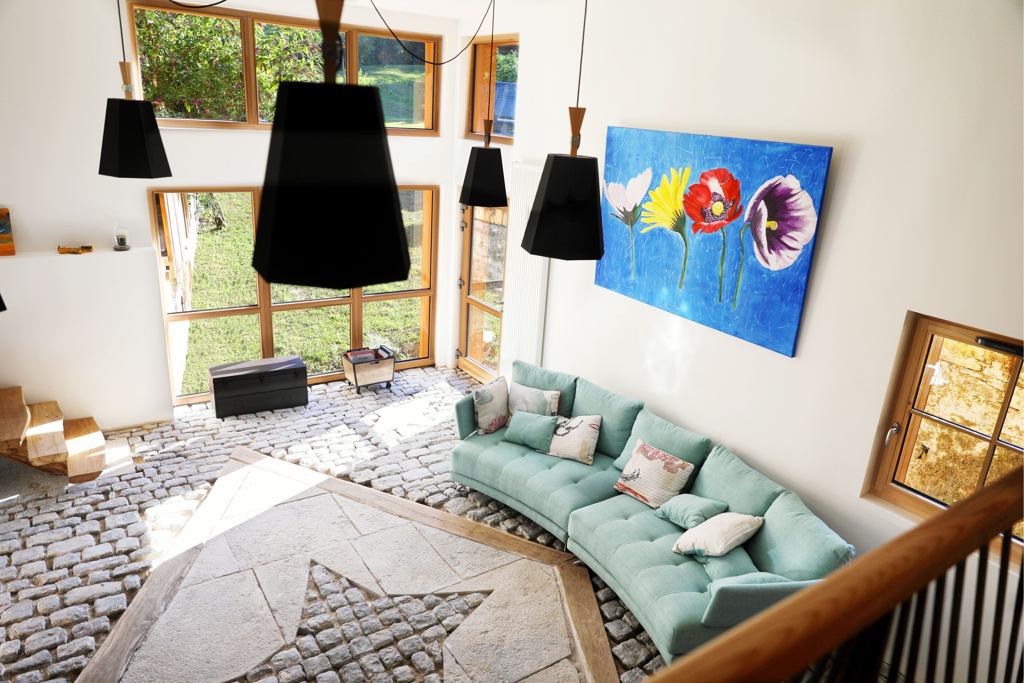 Een moderne woonkamer met een gebogen teal bank, zwarte hanglampen en een groot schilderij van bloemen aan de muur.