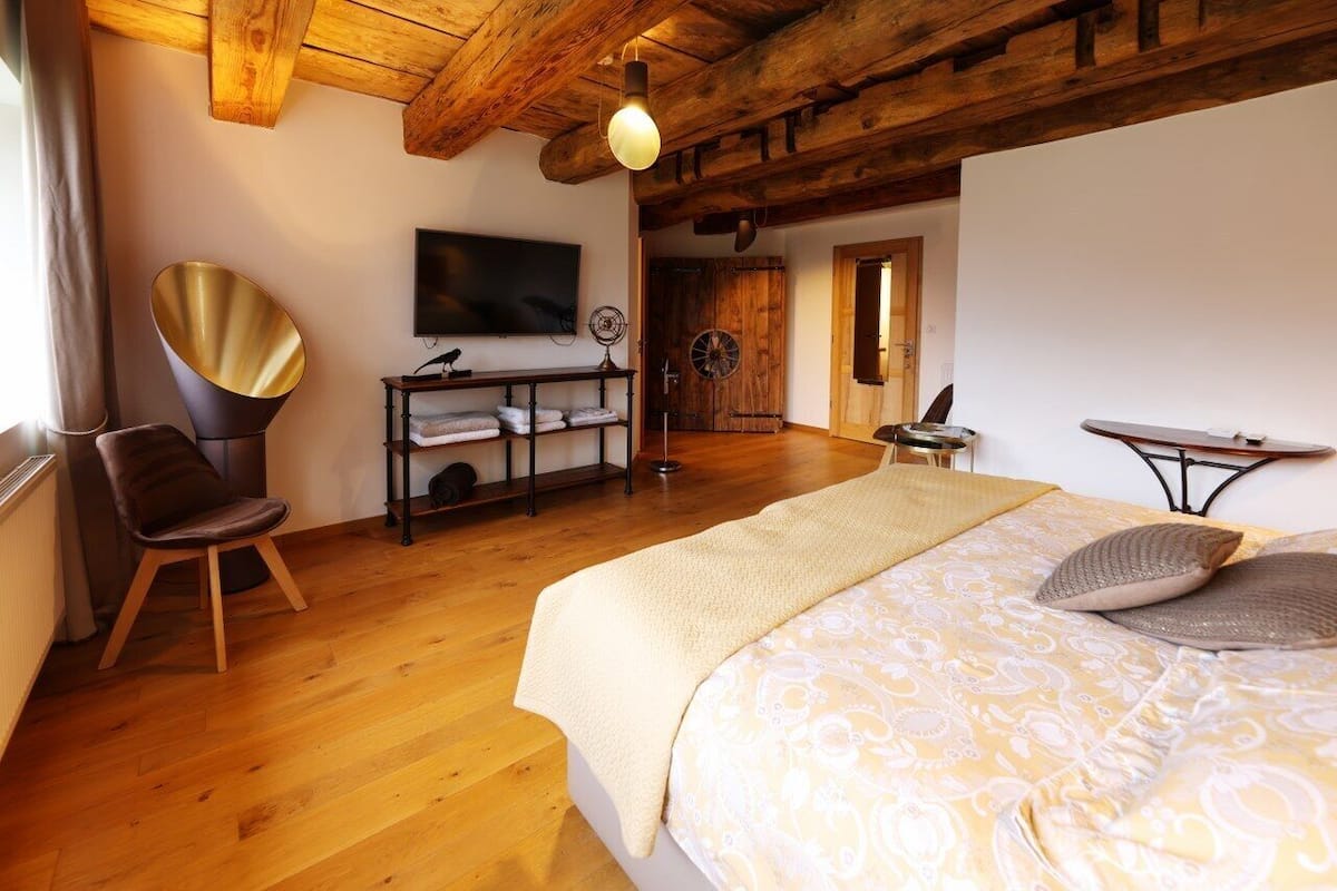 A bedroom with a wooden ceiling, a bed with a patterned duvet, a chair, a desk with a lamp, and a television on the wall.