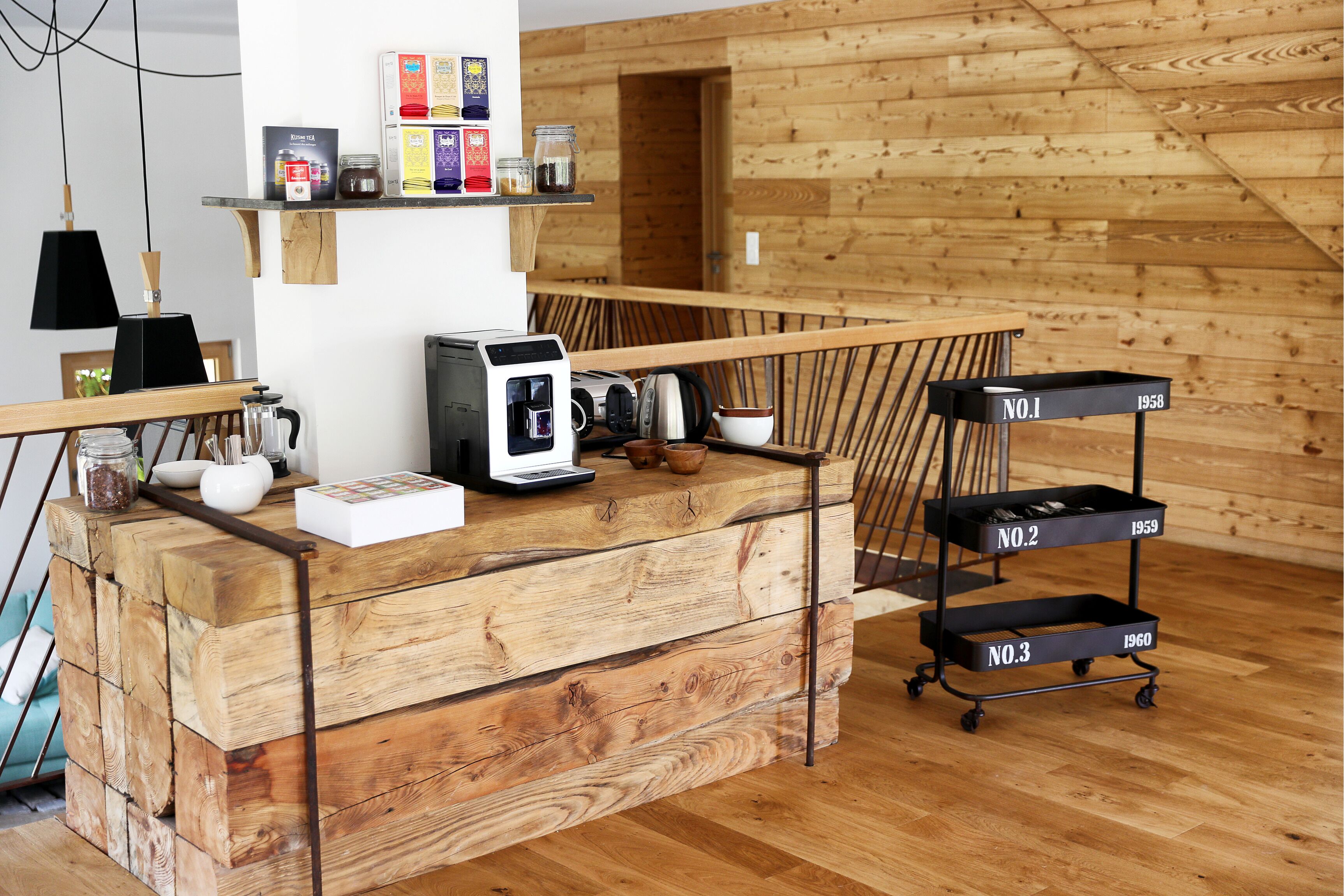 Un comptoir d’accueil en bois, avec une machine à café, une étagère remplie de bocaux et un chariot noir doté de compartiments numérotés.