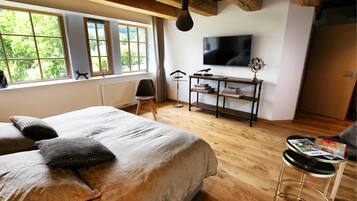 A bedroom with a large bed, a desk with a flat-screen TV, and a window with a view of greenery.