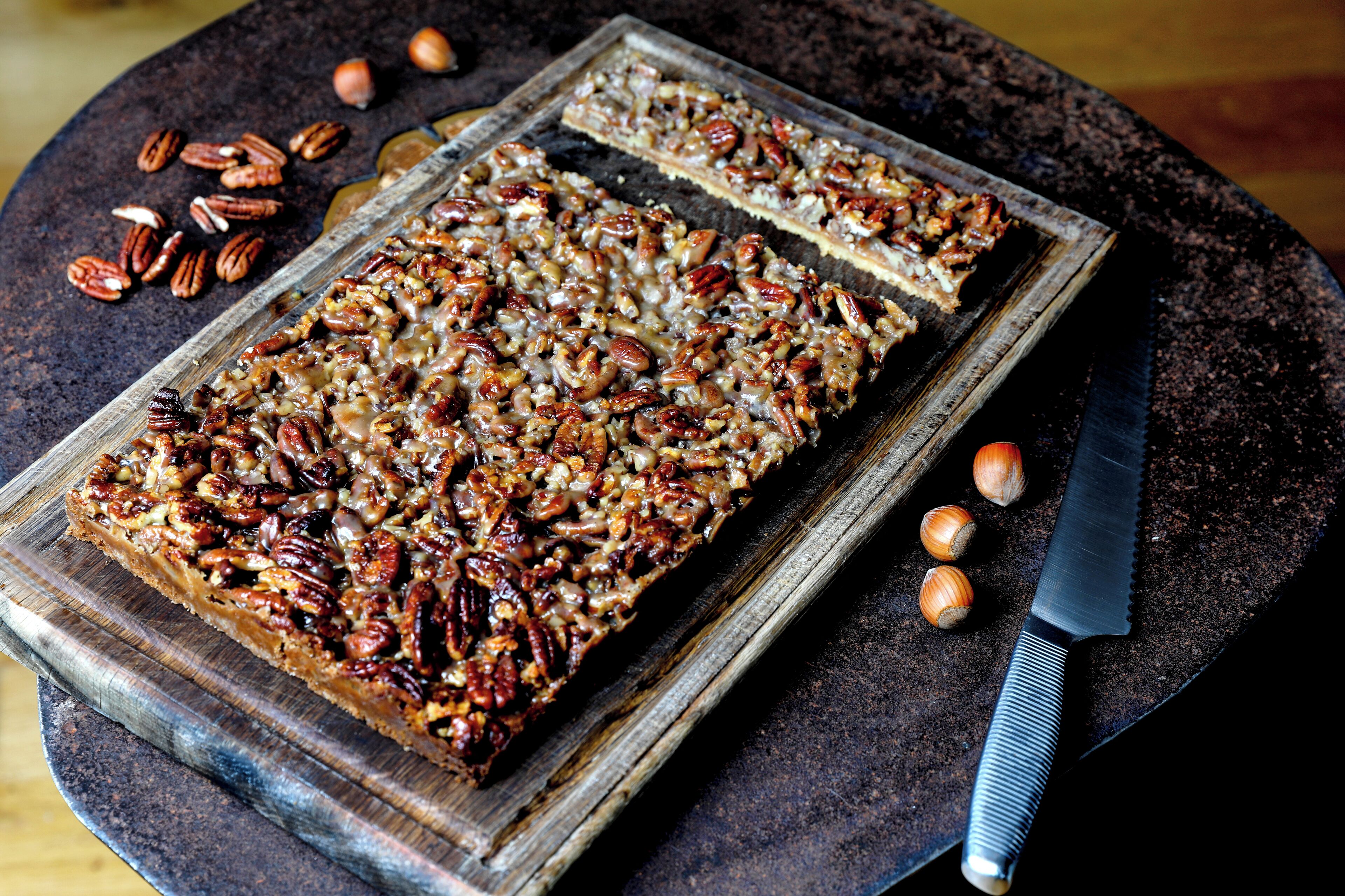 A rectangular tray of pecan-topped dessert on a rustic wooden board, accompanied by a knife and pecans scattered around.
