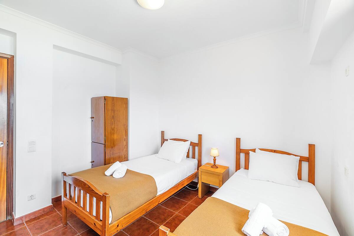A hotel room with two wooden beds, a wooden wardrobe, a nightstand with a lamp, and a tiled floor.