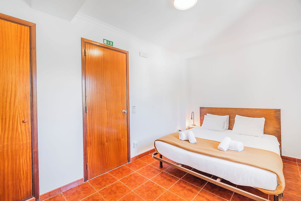 A hotel room with a bed, a wooden door, and a small table with a lamp.