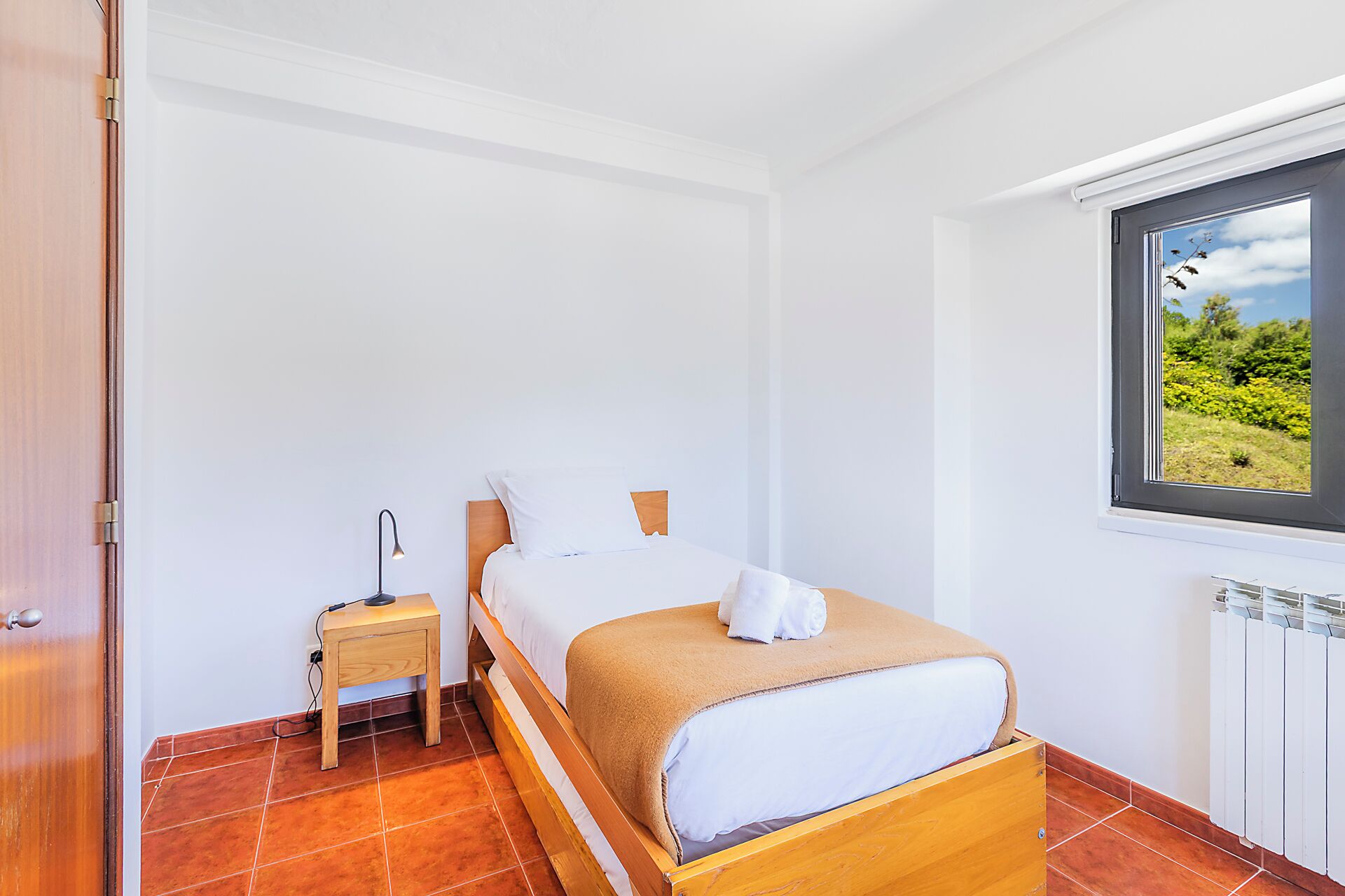 A bedroom with a bed, a nightstand, a window with a view of greenery, and a radiator.