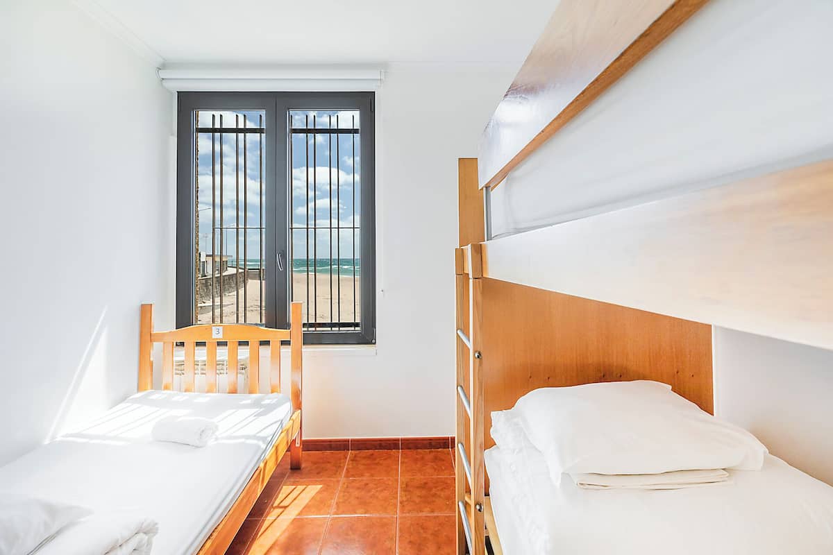 A small, clean room with a single bed, a wooden headboard, a window with a view of the beach, and a tiled floor.
