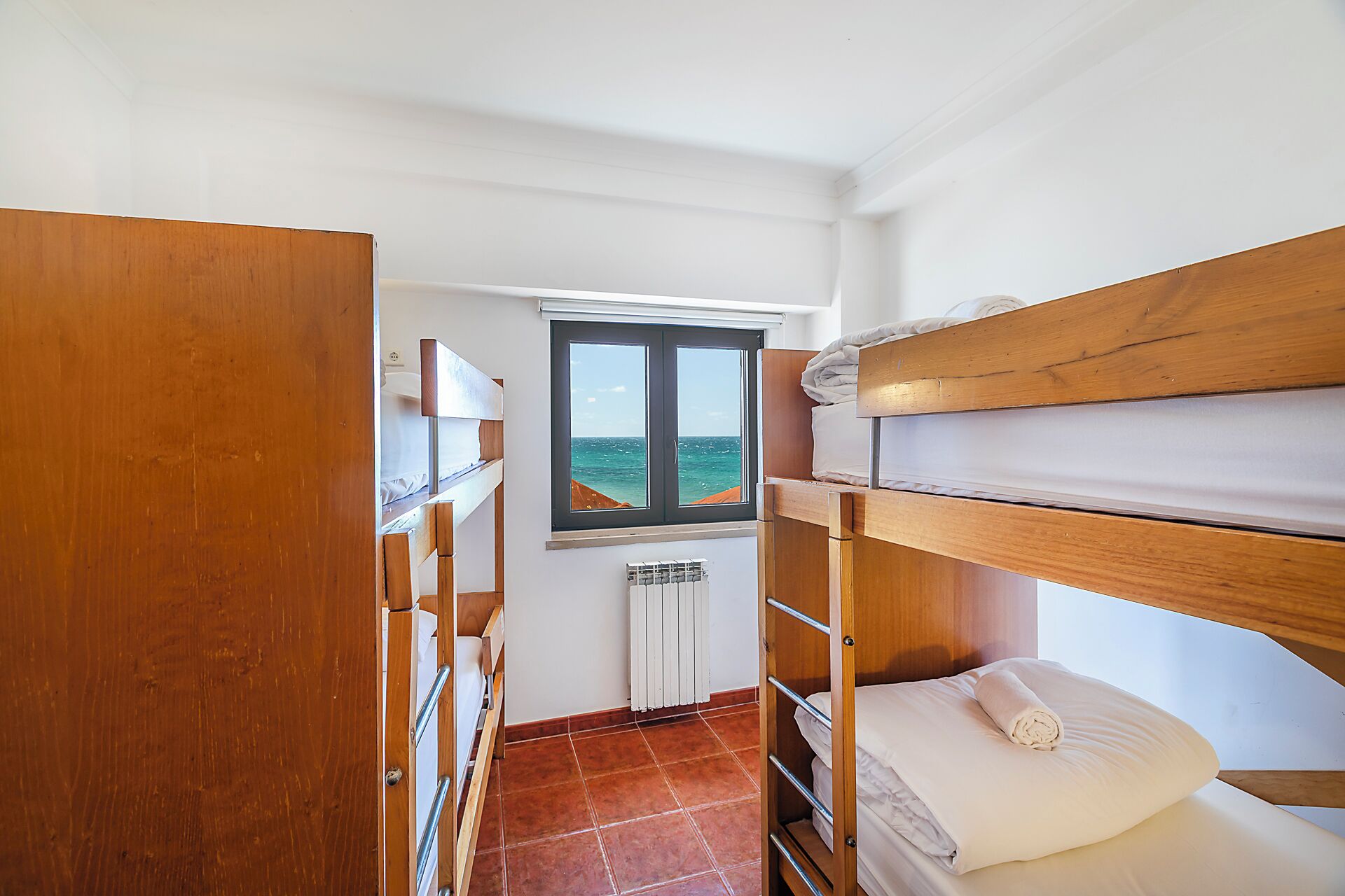Une petite pièce avec des lits superposés en bois, une fenêtre donnant sur la mer et un radiateur posé sur le sol.