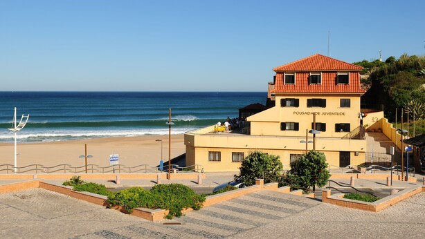 A building named "Pousada Juvenilde" with a red roof, located near a beach with a clear view of the ocean. - HI Areia Branca – Pousada de Juventude - Hostel (Lourinha)