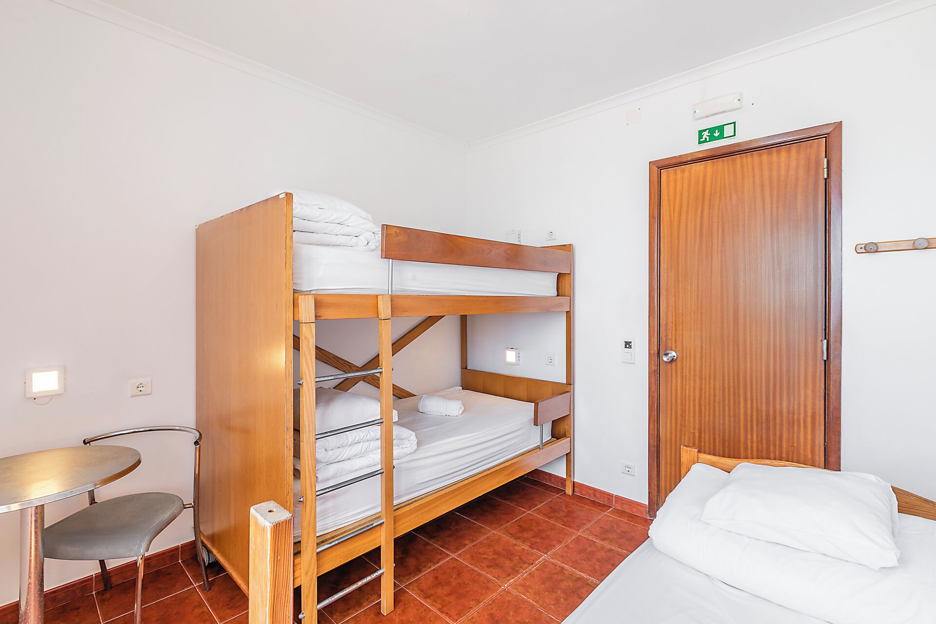 A bunk bed room with a desk and chair, a wooden door, and an exit sign.