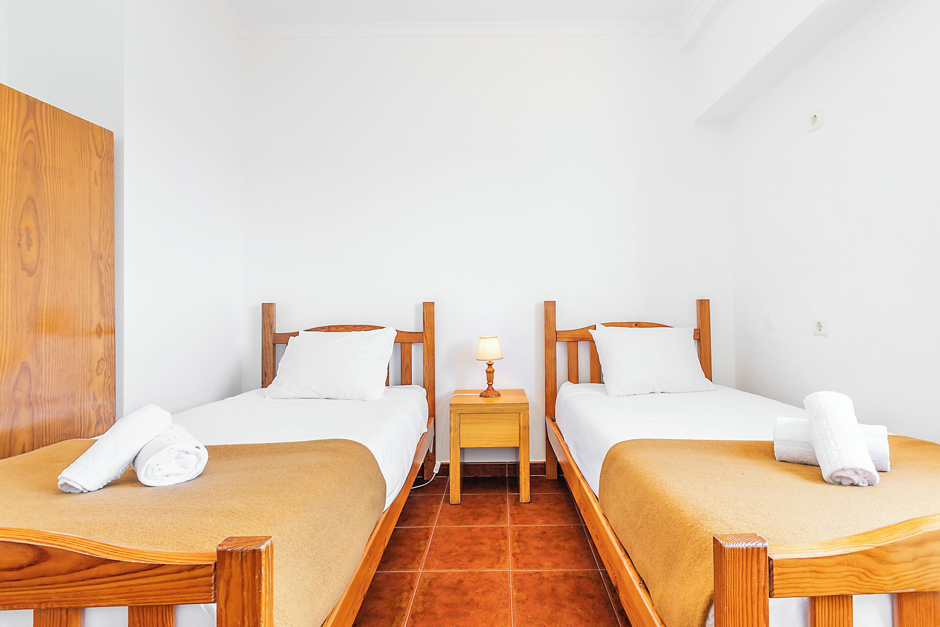 Una camera d'albergo con due letti, testiere in legno, biancheria da letto bianca, un piccolo comodino con una lampada e un armadio in legno.