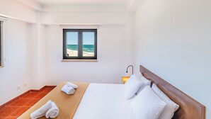 A hotel room with a bed, a window with a view of the sea, a bedside table with a lamp, and a tiled floor. - HI Areia Branca – Pousada de Juventude - Hostel (Lourinha)