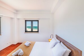 Ein Hotelzimmer mit Bett, Fenster mit Meerblick, Nachttisch mit Lampe und einem Handtuch auf dem Boden.