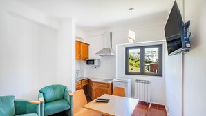 A compact kitchen with wooden cabinets, a microwave, and a window with a view of greenery. - HI Areia Branca – Pousada de Juventude - Hostel (Lourinha)