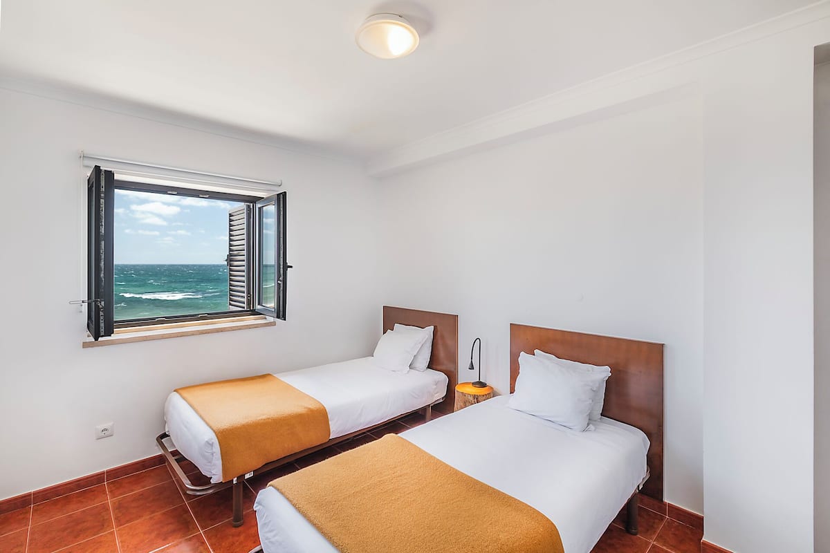 A hotel room with two beds, a window with a view of the sea, and a ceiling light.