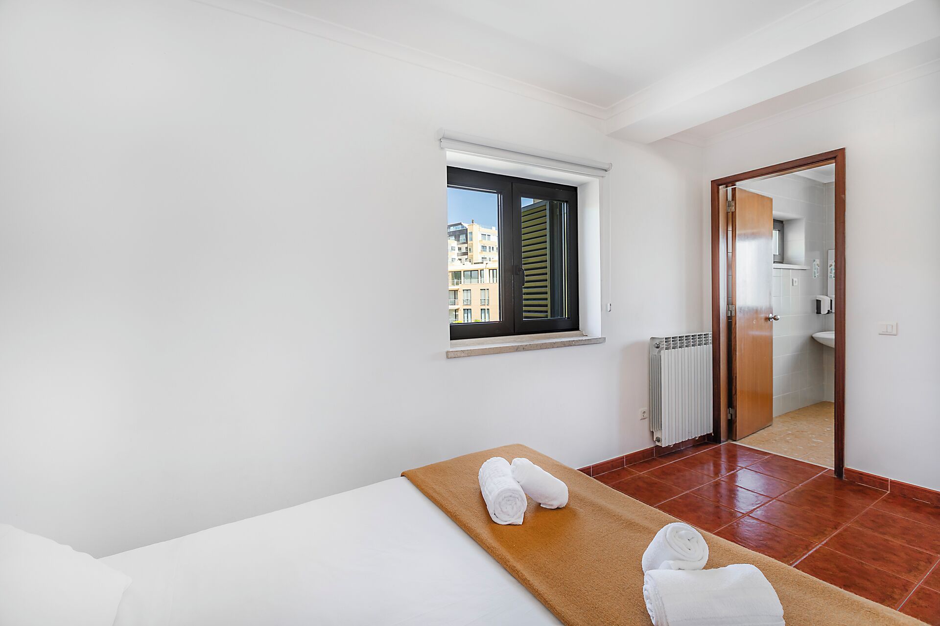 Habitación de hotel con cama, ventana con persianas, radiador y baño visible a través de una puerta abierta.