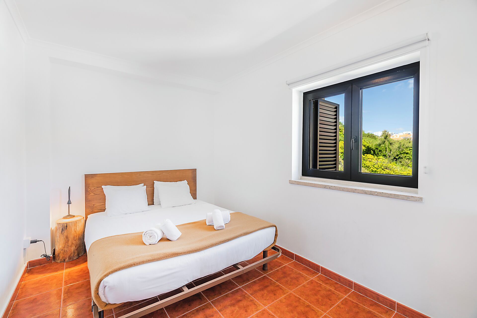 Un dormitorio con cama, ventana con contraventanas, mesita de noche con lámpara y vistas al exterior.