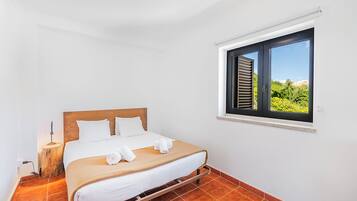 Un dormitorio con cama, ventana con contraventanas, mesita de noche con lámpara y vistas al exterior.