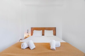 Ein ordentlich bezogenes Bett mit weißen Leinentüchern und zwei aufgerollten Handtüchern auf einer braunen Bettdecke, ein hölzernes Kopfende und eine Nachttischlampe.