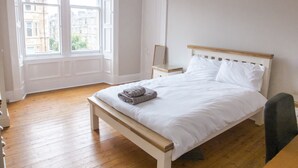Une chambre à coucher avec un lit en bois, une table de chevet, un miroir et une fenêtre donnant sur des immeubles.