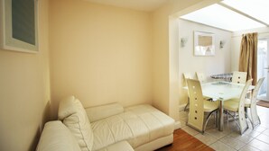 A living room with a beige sofa, a glass-top dining table with chairs, and a view of the outdoors through a window. - Flexistay Hillbrook (London)