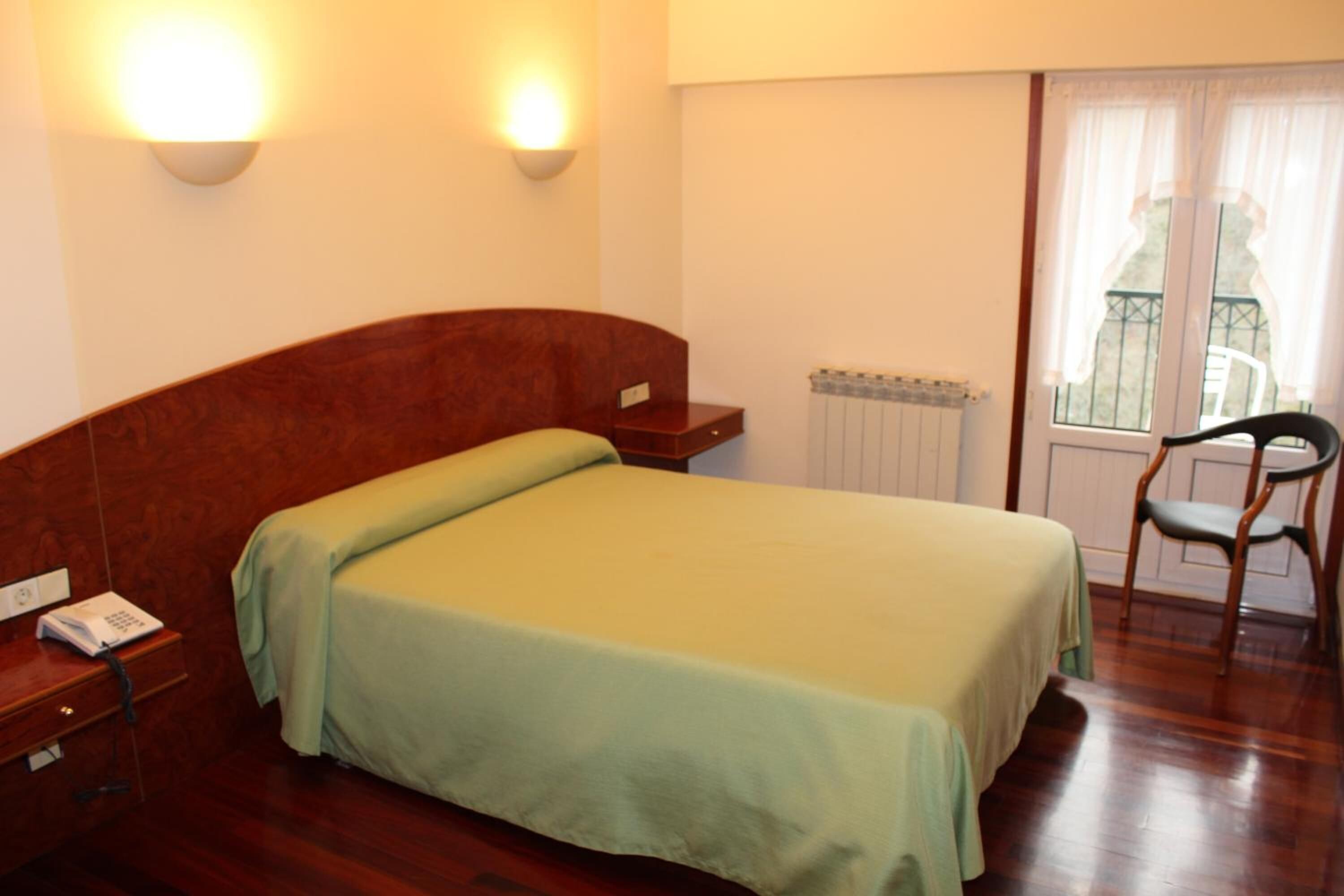 A single-bed room with a wooden headboard, a bedside table with a phone, and a chair near a window with a view.