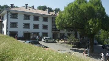 Un immeuble à plusieurs étages, doté d’un toit recouvert de tuiles, avec un grand arbre devant et quelques personnes qui se promènent à proximité.