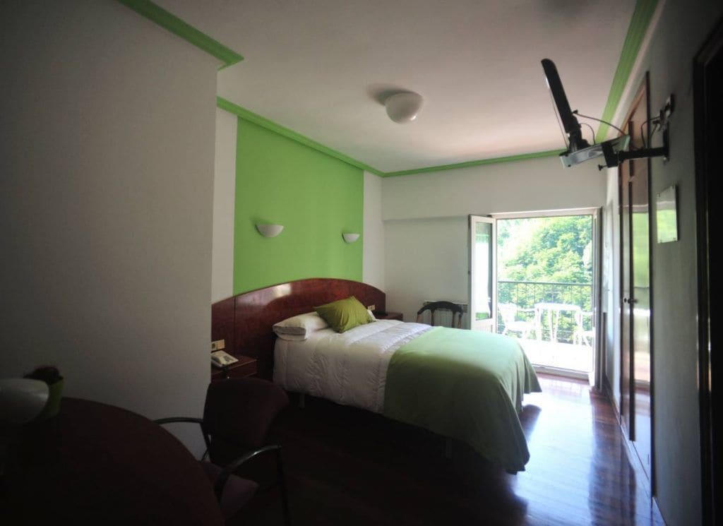 Un dormitorio con cama, escritorio, silla, mesita de noche y ventana con vista a follaje.