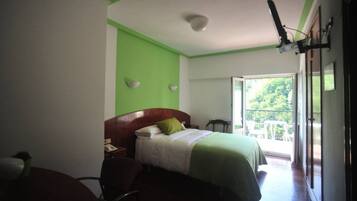 Un dormitorio con cama, escritorio, silla, mesita de noche y ventana con vista a follaje.