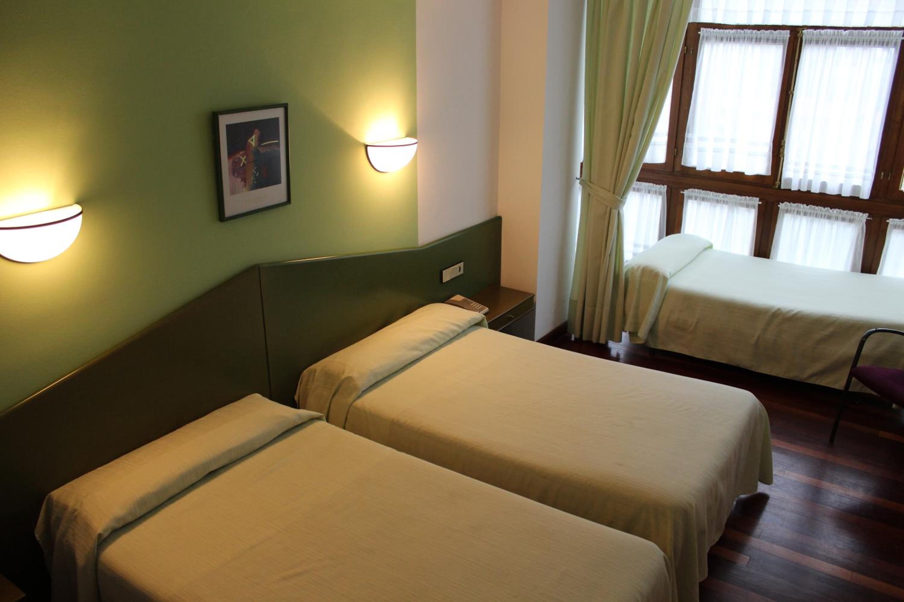 Una habitación con dos camas individuales, una mesita de noche, una lámpara de pared y una ventana con cortinas.